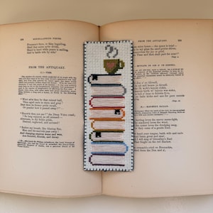 Pu&ograve; includere: Segnalibro fatto a mano con un design a pila di libri e tazza di caff&egrave;. Il segnalibro &egrave; realizzato con la tecnica del punto croce, mostrando vari colori e trame. &Egrave; posto su un libro aperto con testo.