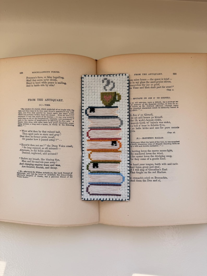 Pu&ograve; includere: Segnalibro fatto a mano con un design a pila di libri e tazza di caff&egrave;. Il segnalibro &egrave; realizzato con la tecnica del punto croce, mostrando vari colori e trame. &Egrave; posto su un libro aperto con testo.