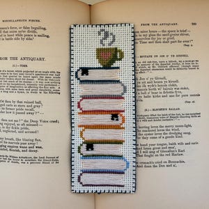 May include: A handmade bookmark featuring a stack of books with colorful spines and a coffee cup on top. The bookmark is made with cross-stitch embroidery and is set against the backdrop of an open book.