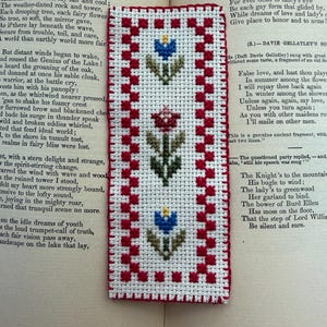 Könnte beinhalten: Handgefertigter Lesezeichen mit weißem Hintergrund und rotem Rand. Das Lesezeichen zeigt drei gestickte Blumendesigns in Blau, Rosa und Grün. Das Lesezeichen liegt auf einem offenen Buch.