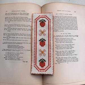 Puede incluir: Un marcapáginas hecho a mano con punto de cruz que presenta un patrón repetido de fresas y flores. El marcapáginas rectangular tiene un borde rojo y está colocado sobre un libro abierto.