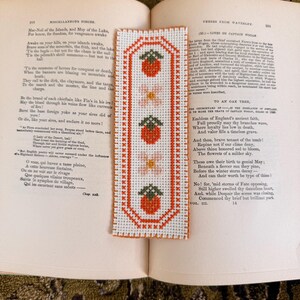 May include: A handmade cross-stitch bookmark featuring a repeating pattern of strawberries and flowers in red, pink, and green on a white background. The bookmark is rectangular with a decorative border and is placed on an open book.