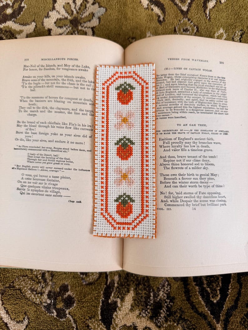 May include: A handmade cross-stitch bookmark featuring a repeating pattern of strawberries and flowers in red, pink, and green on a white background. The bookmark is rectangular with a decorative border and is placed on an open book.