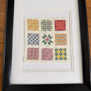 Puede incluir: Obra de arte en punto de cruz enmarcada con nueve cuadrados coloridos con patrones geométricos. Los cuadrados están dispuestos en una cuadrícula de 3x3, con un diseño de estrella central. La obra está enmarcada en un marco negro con un paspartú blanco.