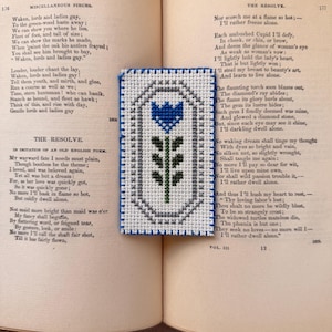 Könnte beinhalten: Handgefertigtes Kreuzstich-Lesezeichen mit einem blauen Blumenmuster und grünem Stiel, vor weißem Hintergrund. Das Lesezeichen liegt auf einem offenen Buch mit Text.