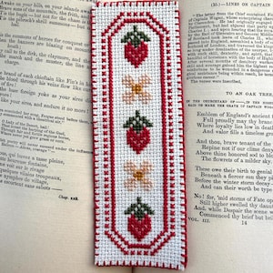 Può includere: Un segnalibro fatto a mano con sfondo bianco e bordo rosso. Il segnalibro presenta un motivo ripetuto di fragole rosse e fiori bianchi. Il segnalibro è posto sopra un libro aperto.