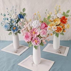 May include: Three bouquets of crocheted flowers in white vases. The flowers are in shades of blue, pink, yellow, and orange, with green leaves. The vases sit on rectangular white cards, arranged on a blue surface.