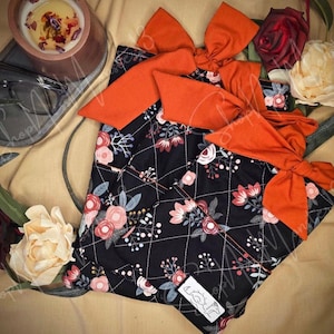 May include: A black floral patterned baby bib with orange bows. The bib features a quilted design with white stitching and a pattern of pink and white flowers and green leaves. The bows are a solid orange color.