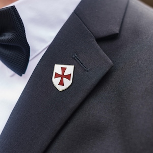 May include: A silver-toned lapel pin featuring a white shield with a red cross design. The pin is attached to a dark navy blue suit jacket. A black bow tie and white dress shirt are also visible.