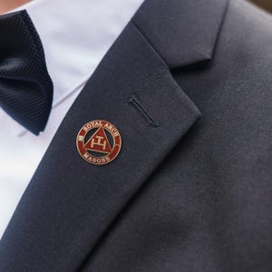 May include: A close-up of a lapel pin with the words "Royal Arch Masons" encircling a red triangle with a gold border. The pin is on a dark navy suit jacket, with a white dress shirt and black bow tie visible.