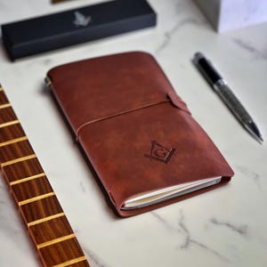 Peut inclure: Un carnet en cuir marron avec une boussole et la lettre G en relief sur la couverture. Le carnet est doté d'une fermeture à sangle et repose sur une surface en marbre blanc.
