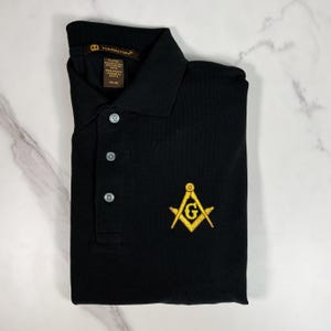 May include: Black polo shirt with a gold embroidered Masonic symbol on the left chest. The shirt has a button-down collar and a three-button placket.