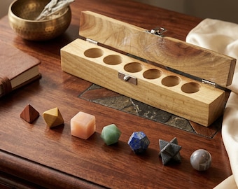 May include: A wooden box with six circular compartments, open to reveal the interior. Various colorful gemstone shapes are arranged in front of the box, including a pyramid, a cube, and a sphere. A brass bowl and a small book are also visible.
