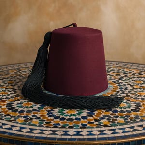 May include: A claret-coloured fez hat with a black tassel. The hat has a cylindrical shape with a flat top and is made of a felt-like material. The black tassel hangs down the side. The hat rests on a mosaic table.