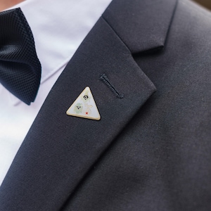 May include: A triangular lapel pin with a white background and colorful floral and geometric designs. The pin is attached to a dark gray suit jacket, near a white collared shirt and a black bow tie.