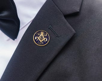 May include: A gold and blue Masonic lapel pin is affixed to a dark blue suit jacket. The pin features a compass, square, and the letter "G" within a circle. A black bow tie and white dress shirt are visible.
