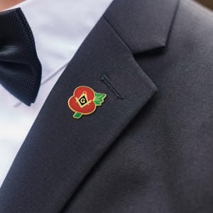 May include: A red poppy pin with a gold outline and a Masonic symbol, worn on a dark navy suit jacket. The pin features green leaves and a gold Masonic symbol in the center. A black bow tie and white shirt are visible in the background.