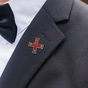 May include: A red and gold cross pin, featuring the letters H, I, S, and V, is pinned to a dark navy suit jacket. The pin is positioned near the lapel, next to a white dress shirt and a black bow tie.