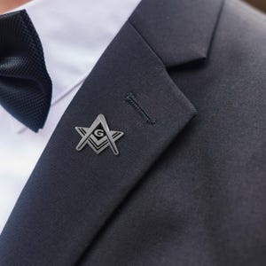 May include: A silver-colored Masonic lapel pin, featuring the Square and Compasses symbol within a star, is affixed to a dark navy suit jacket. A black bow tie and white dress shirt are visible in the background. The pin is a small, detailed accessory.