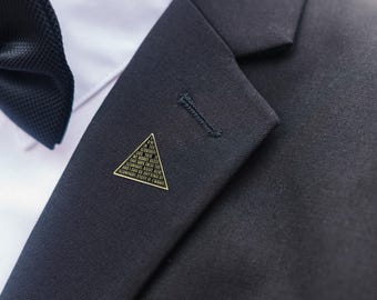 May include: A gold-colored triangular lapel pin with engraved text, affixed to a dark navy suit jacket. The pin is positioned near the lapel buttonhole, next to a white dress shirt and a black bow tie. The text on the pin is legible.