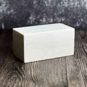 May include: A rectangular, white block with a smooth surface. The block is positioned on a dark wood-grain surface, with a blurred gray background. The block's shape is a simple, solid form.