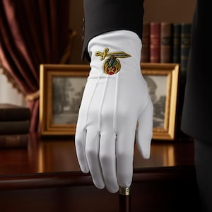 May include: White dress glove with an embroidered emblem featuring a sword and floral design in gold, red, and green. The glove is held over a dark wooden surface, suggesting formal attire or ceremonial use.