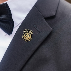Pode incluir: Um alfinete de distintivo da polícia dourado e preto com o símbolo maçónico, preso a um casaco de fato azul marinho escuro. O alfinete apresenta a palavra "POLÍCIA" e um emblema maçónico. Uma gravata borboleta preta é visível.