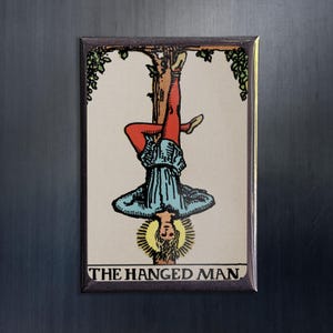 Könnte beinhalten: Eine Tarotkarte mit einer bunten Illustration eines Mannes, der kopfüber von einem Ast hängt. Die Karte trägt den Titel "Der Gehängte".