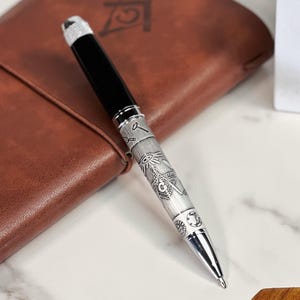Può includere: Una penna d'argento con un cappuccio nero e un design massonico. La penna è incisa con un compasso, una squadra e un occhio che tutto vede.