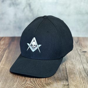 Op de afbeelding: Een zwarte baseballpet met een wit geborduurd schedel en gekruiste beenderen in een kompasontwerp.