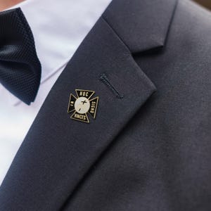 May include: A gold and black lapel pin, inscribed with "HOC IN SIGNO VINCES", is pinned to a dark navy blue suit jacket. The pin is cross-shaped with a white centre and a cross symbol. A black bow tie and white shirt are also visible.