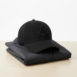 Könnte beinhalten: Eine schwarze Baseballkappe mit einem Freimaurersymbol, die auf einem gefalteten dunkelgrauen Anzugsakko liegt. Die Kappe zeigt ein erhabenes Emblem aus Winkel und Zirkel mit dem Buchstaben "G" in der Mitte. Das Sakko ist ordentlich auf einer hellen Holzoberfläche gefaltet.