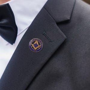 以下が含まれることがあります： フリーメイソンのエンブレムが付いたラペルピンのクローズアップ。 ピンは丸く、青い縁と金色のディテールがあります。 「FREE AND ACCEPTED MASONS」の文字がエンブレムの周りに刻まれています。 ピンは濃い色のスーツのジャケットに取り付けられています。