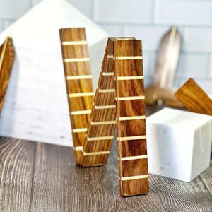 Puede incluir: Una escultura de madera con forma de la letra "N" con un acabado marrón cálido y rayas incrustadas de color claro. La escultura está hecha de madera y tiene un diseño geométrico. El fondo incluye una forma geométrica blanca y un cubo blanco.