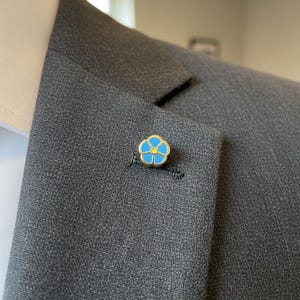 May include: A gold-toned flower lapel pin with a blue and yellow enamel design. The pin is attached to a gray suit jacket, near the lapel buttonhole. The flower has five petals.