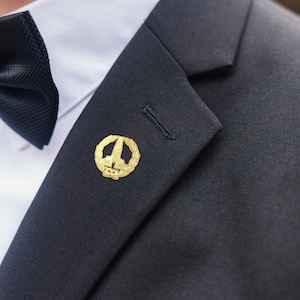 Könnte beinhalten: Goldfarbene Anstecknadel mit detailliertem Design, mit einem zentralen Emblem, das von einem Lorbeerkranz umgeben ist. Die Nadel ist an einem dunkelblauen Anzugsakko befestigt, kombiniert mit einem weißen Hemd und einer schwarzen Fliege.