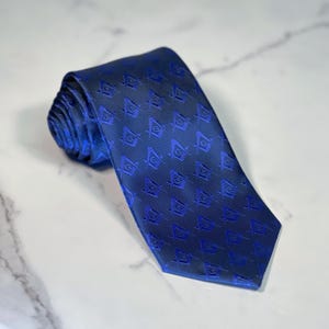 Könnte beinhalten: Eine gerollte marineblaue Krawatte mit einem sich wiederholenden Muster aus Freimaurersymbolen in einem helleren Blauton. Der Stoff der Krawatte hat einen dezenten Glanz und wird auf einer Marmoroberfläche präsentiert. Das Design ist formell und elegant.