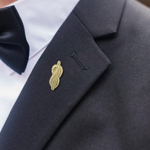 Pode incluir: Um alfinete de lapela dourado em forma de pena estilizada está preso a um blazer azul marinho escuro. O alfinete está posicionado perto da casa do botão da lapela. Uma gravata borboleta preta e uma camisa branca são visíveis no fundo.