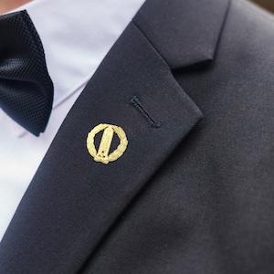 Könnte beinhalten: Goldfarbene Anstecknadel mit einem zentralen Design, umgeben von einem Lorbeerkranz, an einem dunkelblauen Anzug befestigt. Der Anzug wird mit einem weißen Hemd und einer schwarzen Fliege getragen. Nahaufnahme.