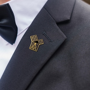 May include: A gold Masonic lapel pin, featuring a square and compass design, is attached to a dark navy suit jacket. The pin is positioned near the lapel buttonhole. A white dress shirt and black bow tie are visible in the background.