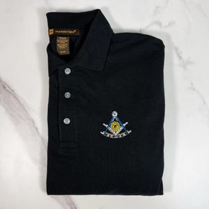 Może przedstawiać: Czarny polo z małym haftowanym symbolem masońskim na lewej piersi. Symbol jest niebieski, biały i żółty i przedstawia cyrkiel i kątomierz.