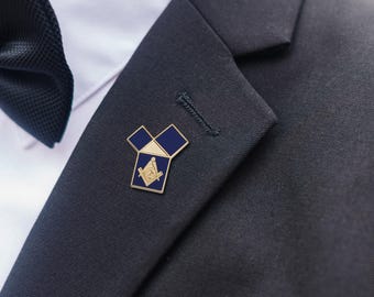 Puede incluir: Un pin de solapa masónico dorado con detalles de esmalte azul y blanco, fijado a una chaqueta de traje azul marino oscuro. El pin presenta el símbolo de la escuadra y el compás, un emblema clave de la masonería. Una pajarita negra y una camisa blanca son visibles.