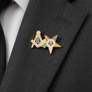 Può includere: Spilla da bavero dorata con simboli massonici. La spilla include un compasso e una squadra con una "G" blu e una stella multicolore. La spilla è attaccata a una giacca da abito grigio scuro.