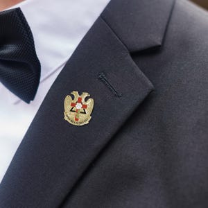 Puede incluir: Un pin de solapa dorado con una cruz roja, una rosa y la inscripción "SPES MEA IN DEO EST" está prendido a un blazer azul marino oscuro. El pin presenta un diseño de águila a ambos lados de la cruz. Una pajarita negra es visible en el fondo.
