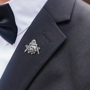 Könnte beinhalten: Silberfarbene Freimaurer-Anstecknadel mit Totenkopf, Zirkel und Winkelmaß-Design. Sie ist an einem dunkelblauen Anzugsakko befestigt, mit einem weißen Hemd und einer schwarzen Fliege im Hintergrund. Ein Symbol der Freimaurerei.