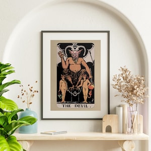 Op de afbeelding: Een vintage tarotkaart-illustratie van de kaart De Duivel. De kaart toont een roodhuidige, gehoornde en gevleugelde demon die op een troon zit. Twee naakte figuren zijn aan de troon geketend. De kaart is in zwart ingelijst en heeft een witte achtergrond.