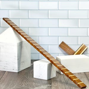 May include: A long, wooden ruler with brass inlay markings rests on a white marble block. The ruler is angled diagonally across the image, with the end of the ruler extending off the right side of the image. The ruler is resting on a white marble block, and there is a second white marble block to the left of the ruler. The background is a white tiled wall.