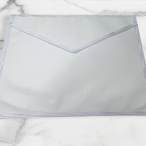 May include: A light gray apron with a pointed top and a rectangular bottom. The apron has a light gray trim around the edges and a light gray trim that forms a V-shape at the top.