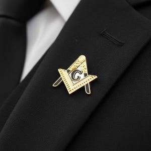 May include: A gold-colored Masonic lapel pin with black and white accents, featuring a compass and square design with the letter "G" in the center. The pin is affixed to a black suit jacket.