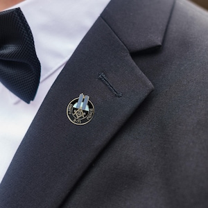 May include: A close-up of a lapel pin with a black border, featuring the Twin Towers, a compass, and the text "UNITY" and "PEACE". The pin is gold-toned and is affixed to a dark navy suit jacket. A black bow tie is visible in the background.
